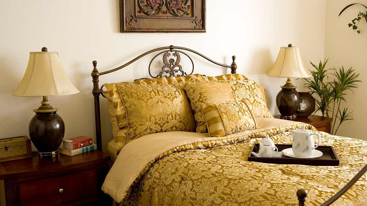 A cozy bedroom with ornate bedding and decorative pillows, featuring a metal headboard and a warm lamp on the bedside table.