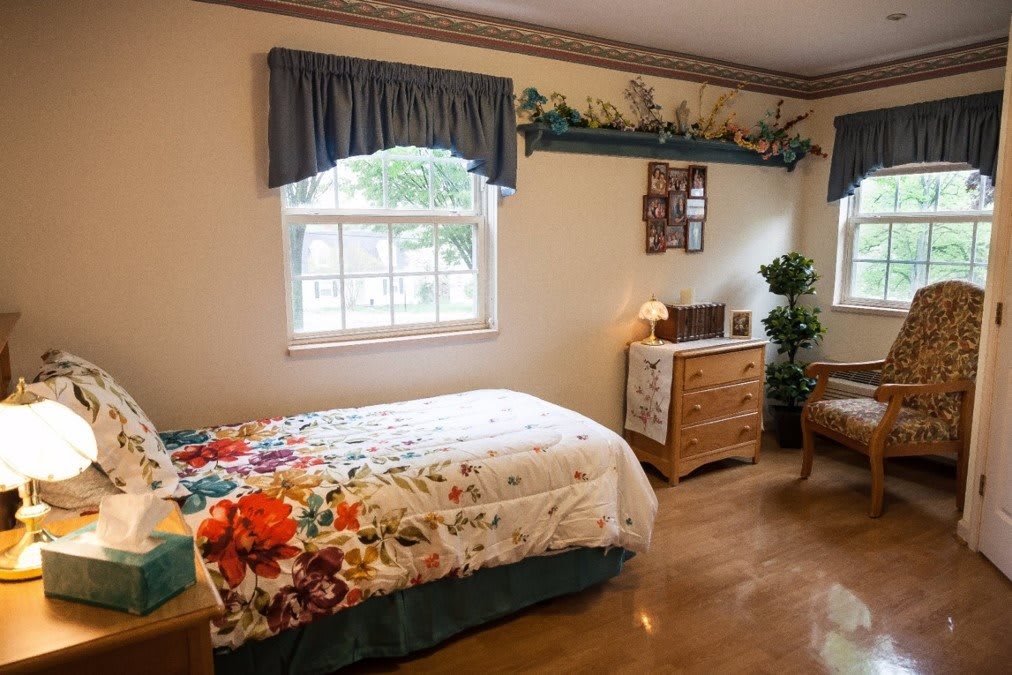 A cozy bedroom featuring a single bed with floral bedding, a window with blue curtains, and wooden furniture.