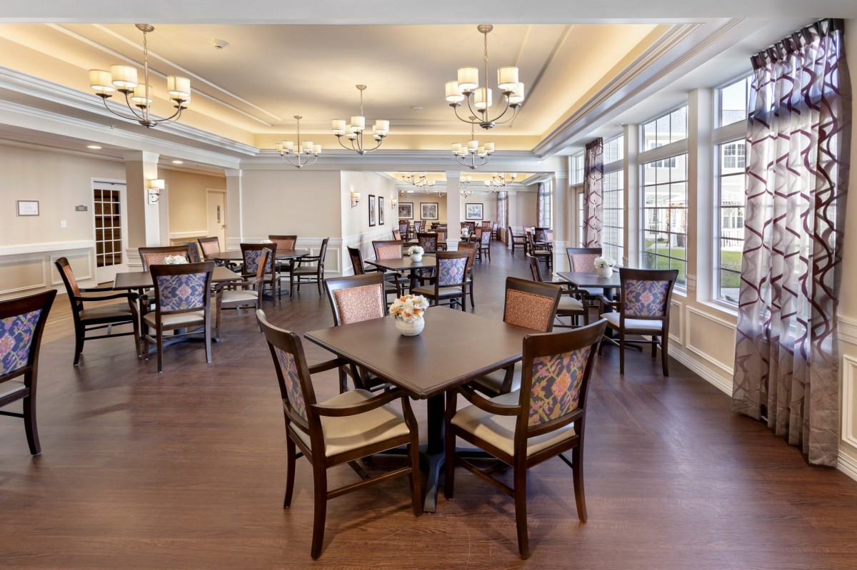 A modern dining room with elegant chandeliers, featuring several tables and chairs set on a wooden floor, surrounded by large windows.