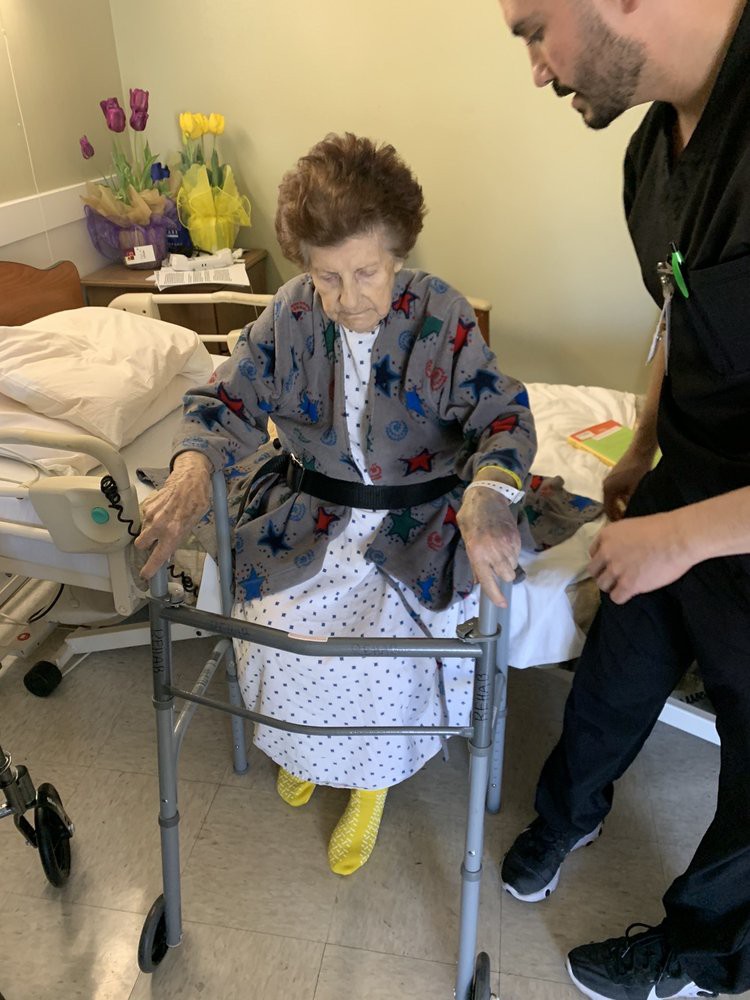 An elderly person using a walker with assistance from a caregiver in a hospital room.