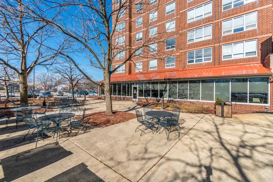 A view of an outdoor patio with tables and chairs in front of a brick apartment building.