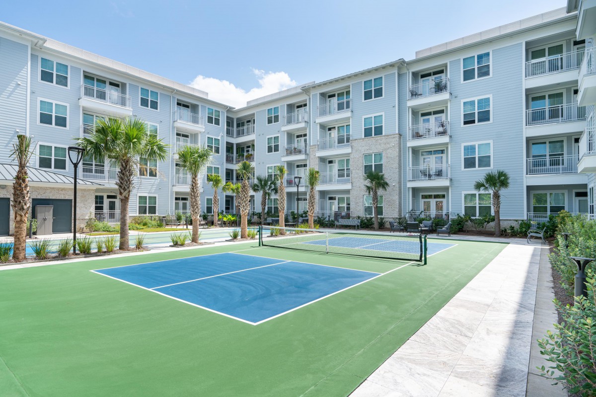 A modern apartment complex with an outdoor tennis court and palm trees under a clear blue sky.