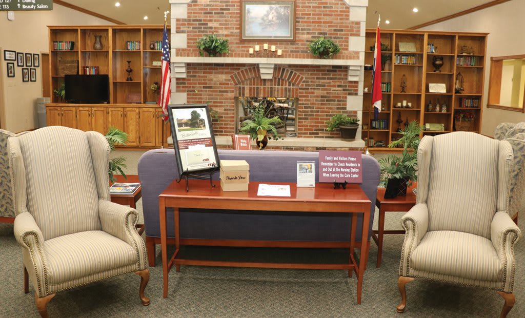 A cozy library lounge area with a sofa, chairs, and a wooden table in front of a brick fireplace, surrounded by bookshelves and decorative plants.