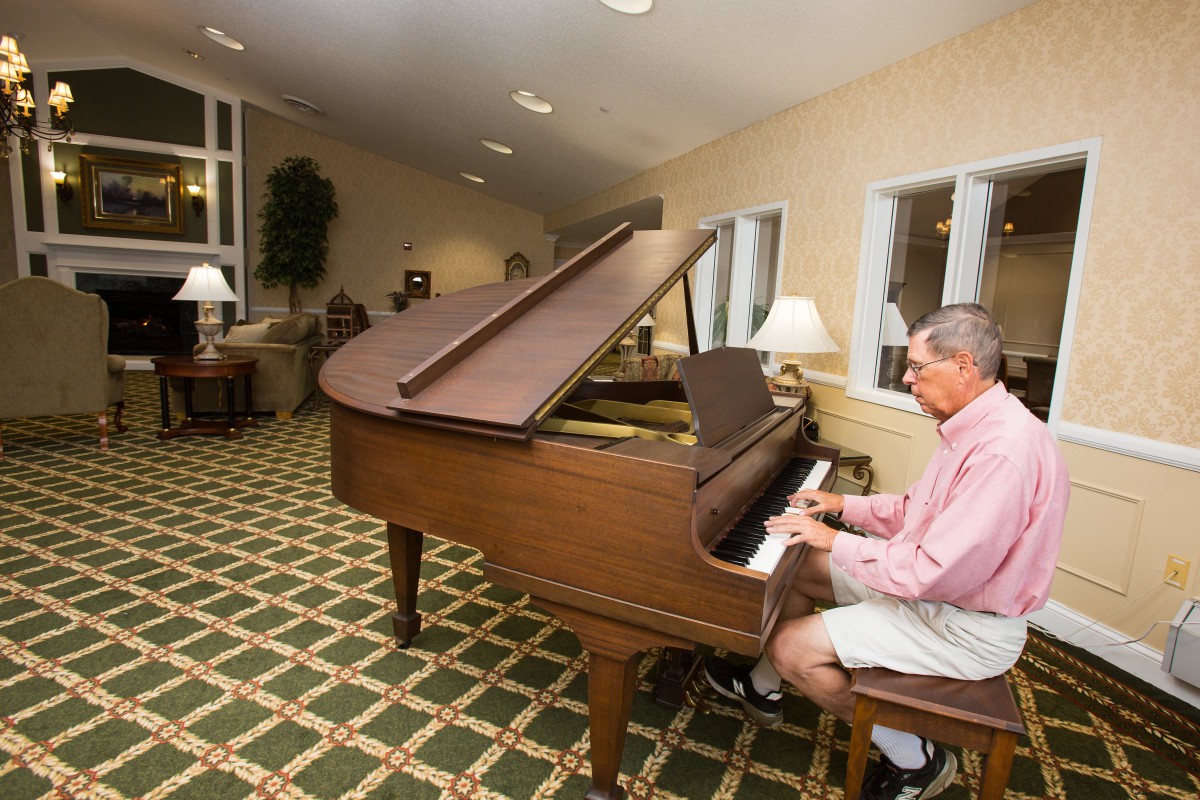 A man playing a grand piano in a room with patterned carpet and furniture.