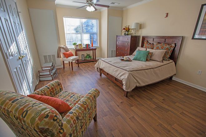 A cozy bedroom with a wooden floor, featuring a bed, chairs, and a large window.