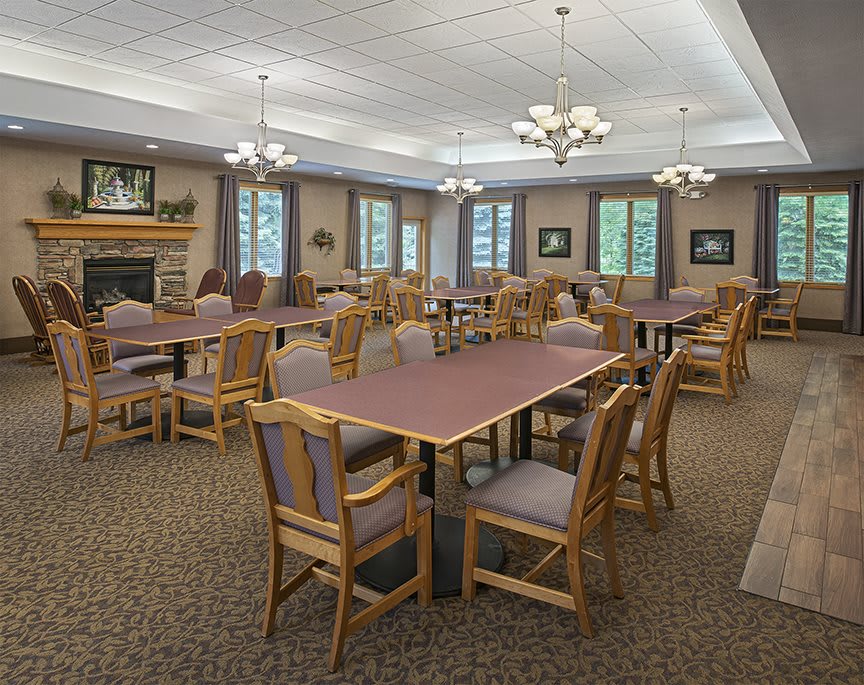A spacious dining room with multiple tables and chairs, well-lit by chandeliers and natural light from the windows.