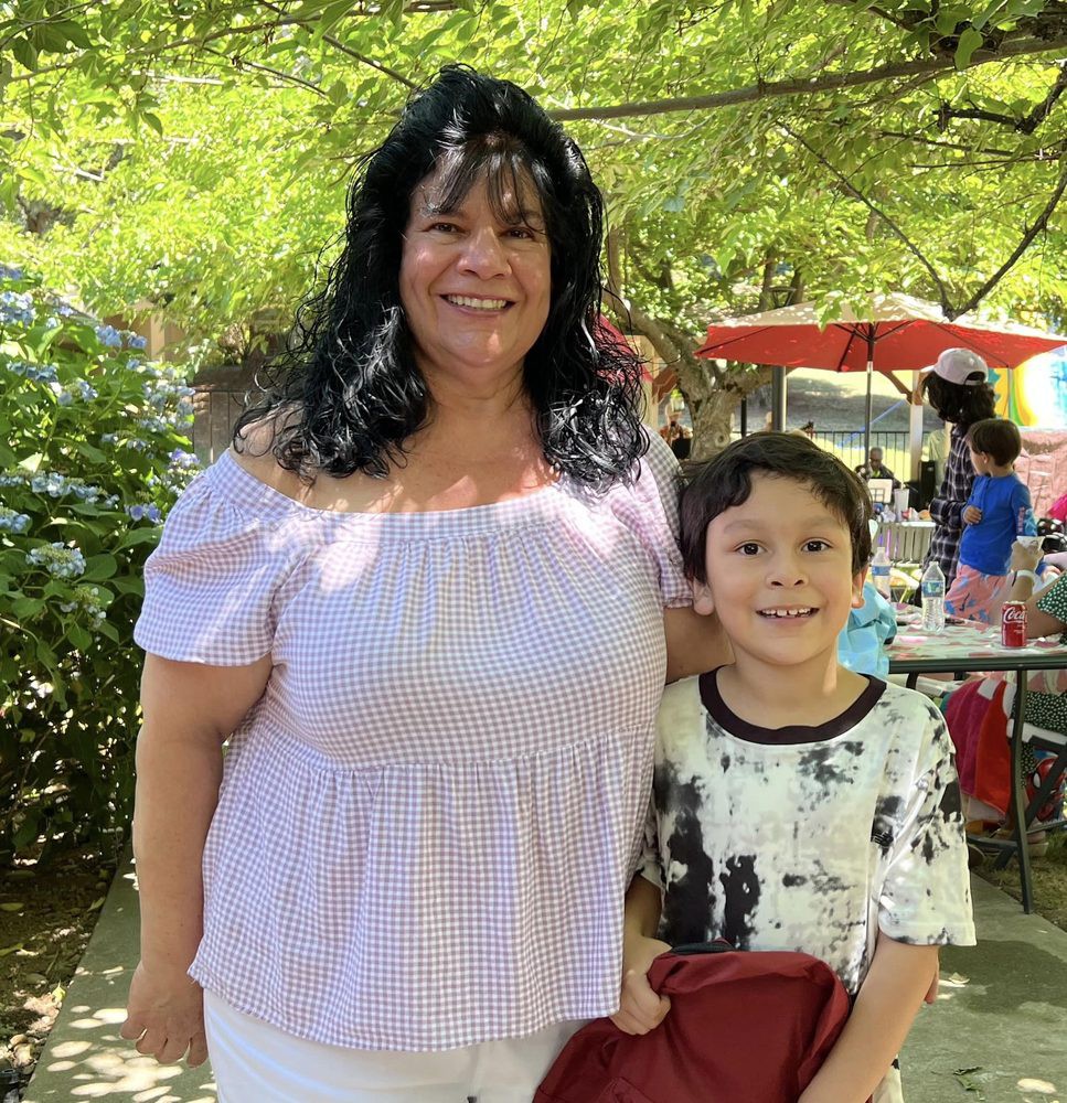 A woman and a child smiling outdoors in a sunny park setting.
