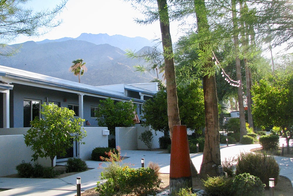 A modern residential building surrounded by trees with mountains in the background on a sunny day.