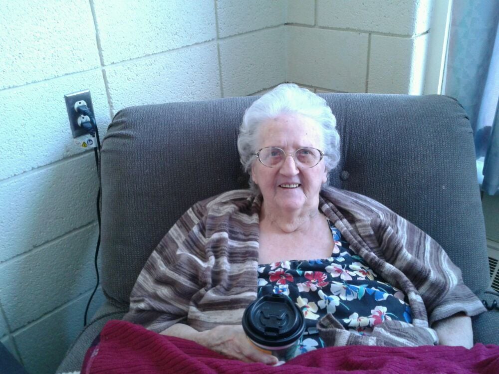 An elderly woman sitting comfortably in a chair, smiling while holding a coffee cup.
