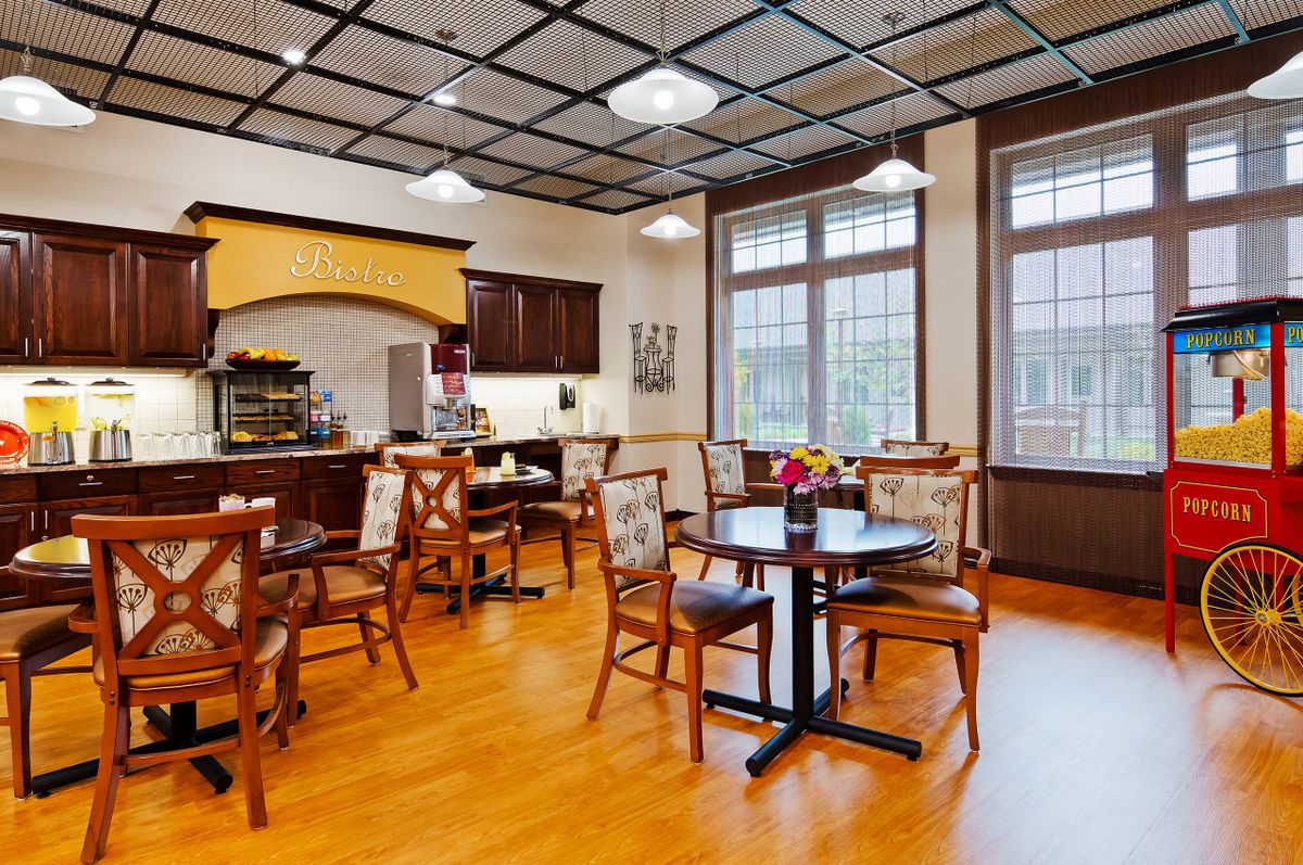 A cozy cafe interior with wooden flooring, tables, and chairs, illuminated by ceiling lights and natural light from large windows.