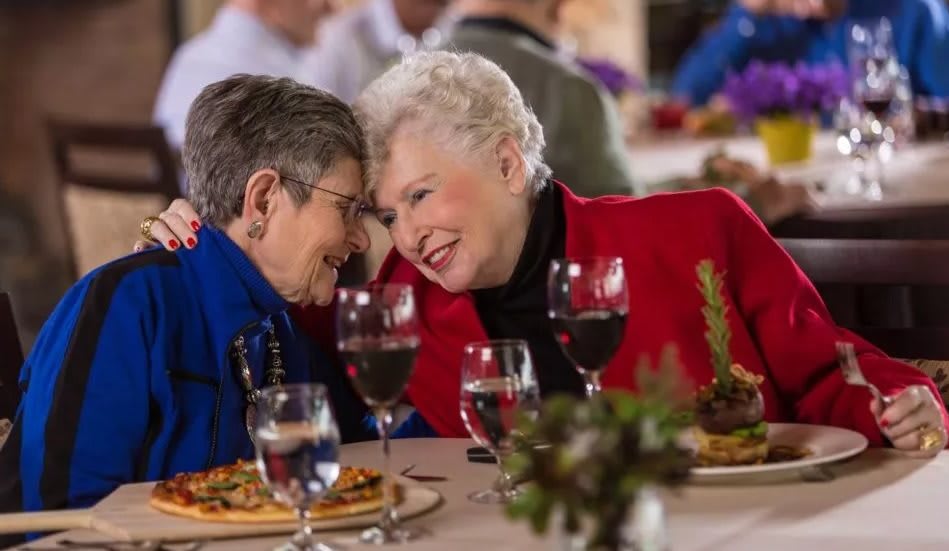 Two elderly women share a warm moment at a dinner gathering, surrounded by plates of food and glasses of wine.
