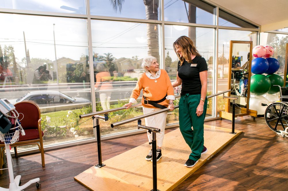An elderly person receives assistance from a healthcare professional while walking between parallel bars for rehabilitation.