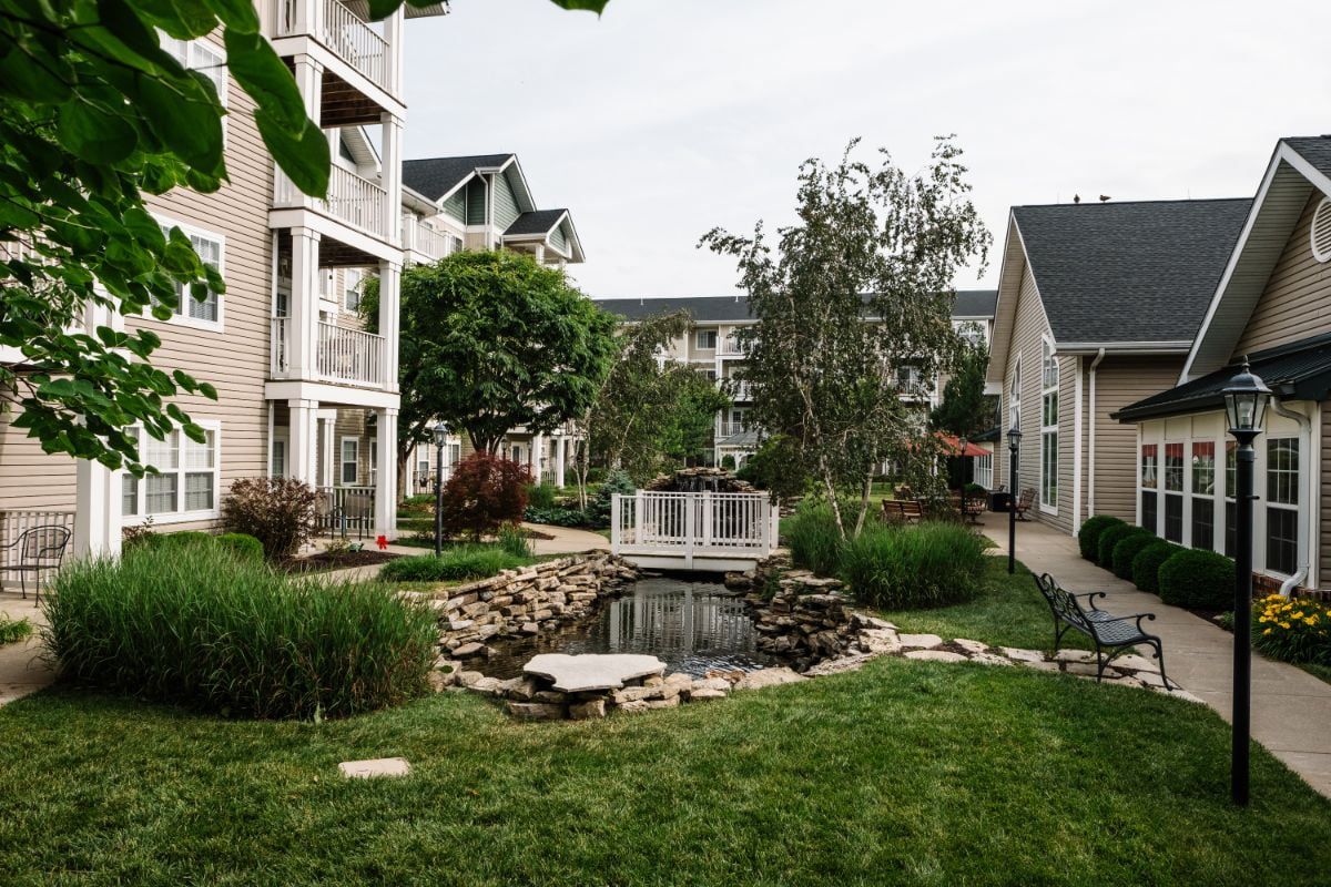 A serene apartment complex with lush greenery, featuring a small pond and bridge.
