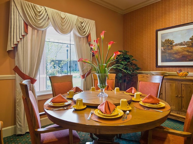 A well-decorated dining room featuring a round wooden table set with orange napkins and yellow dishes, complemented by floral arrangements and elegant curtains.
