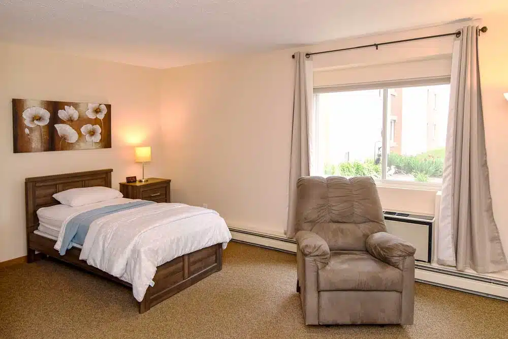 A cozy bedroom with a bed, recliner chair, and a window letting in natural light.