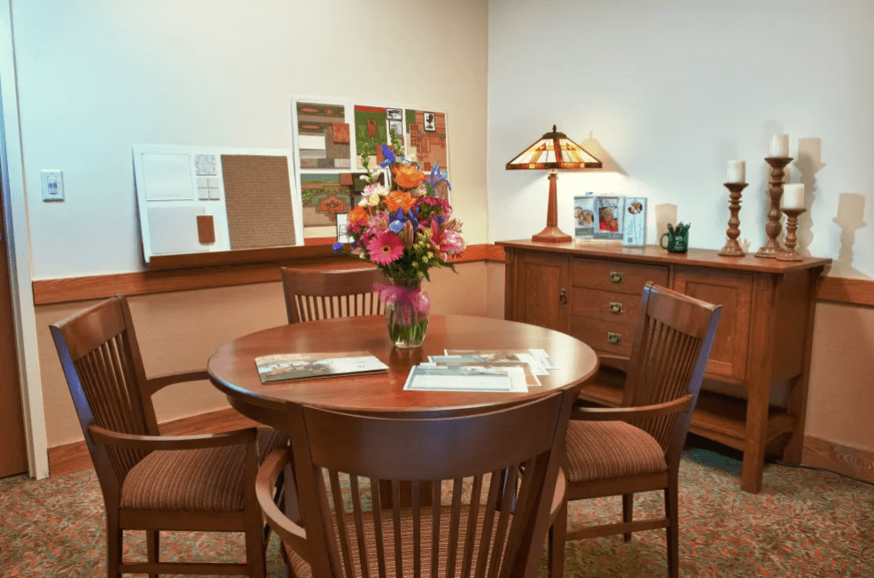 A cozy office meeting room with a round wooden table, four chairs, and a vase of flowers.