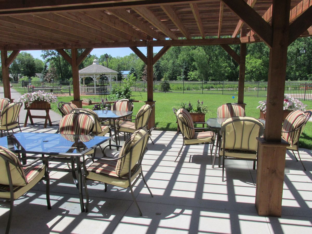 A sunny outdoor patio with tables and chairs under a pergola, overlooking a garden.
