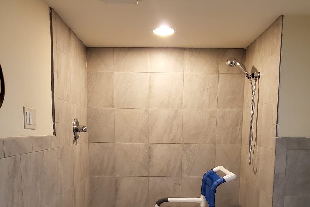 A tiled bathroom featuring a shower area with a shower chair under bright lighting.