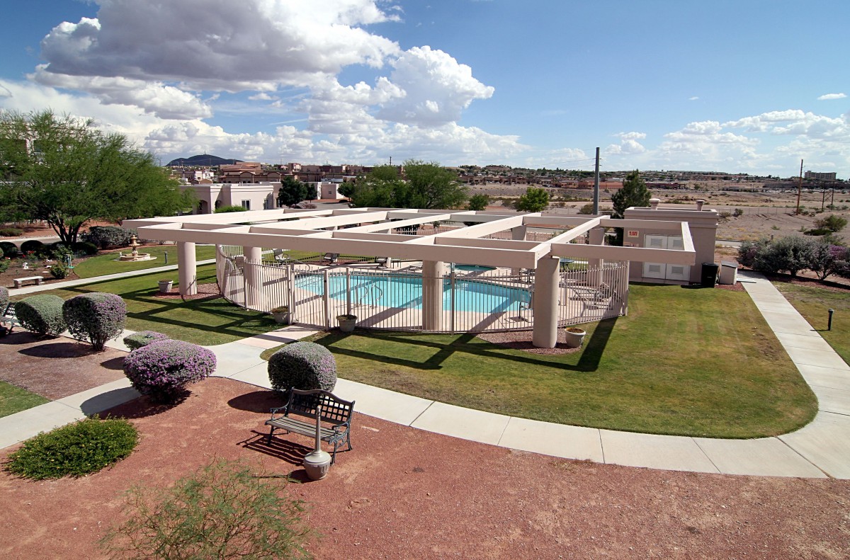 A scenic view of an outdoor swimming pool area with a pergola, surrounded by grass and under a partly cloudy sky.