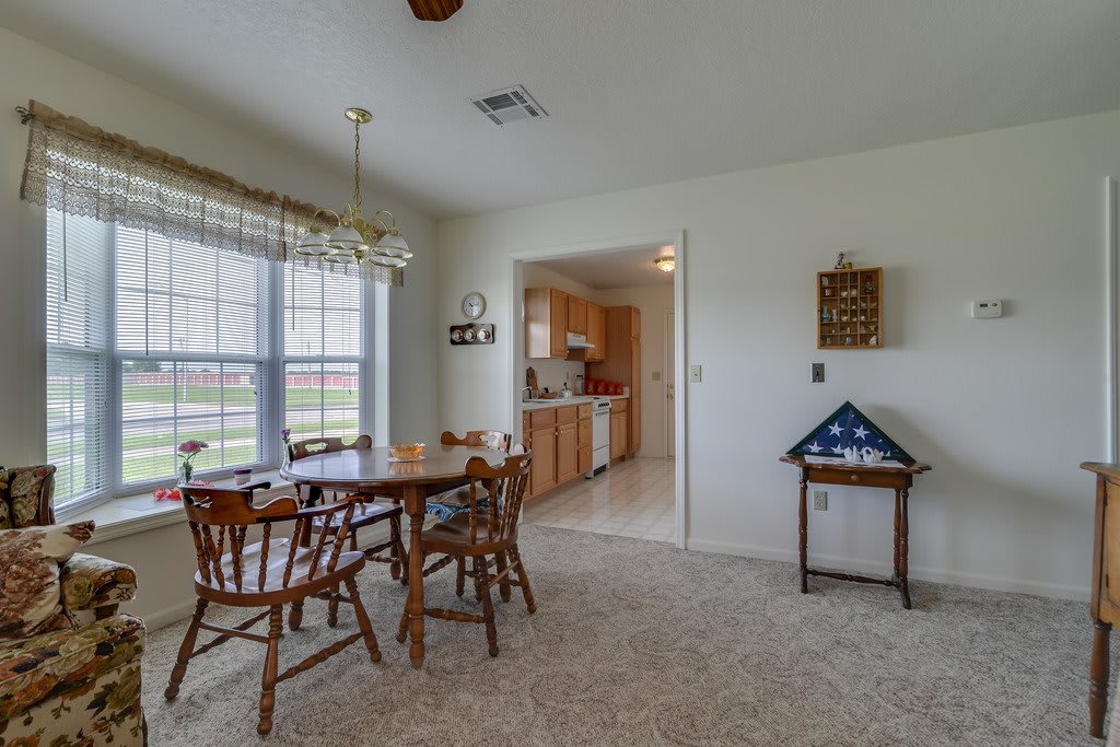 A cozy dining area with a wooden table and chairs, adjacent to a kitchen, featuring large windows and carpet flooring.