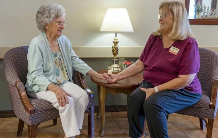An elderly person and a caregiver hold hands while sitting together indoors.