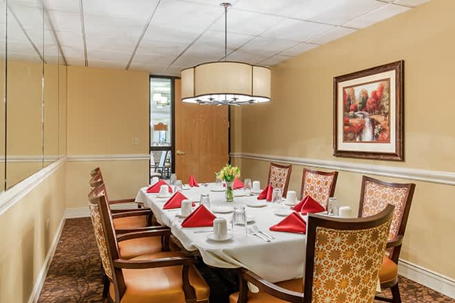 A formal dining room with a neatly set table, featuring red napkins, elegant chairs, and a modern chandelier.