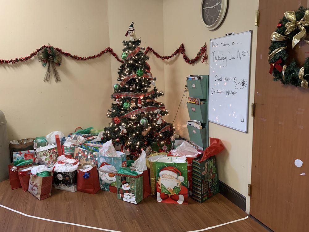 A festive office corner with a decorated Christmas tree surrounded by wrapped gifts and a whiteboard displaying event information.