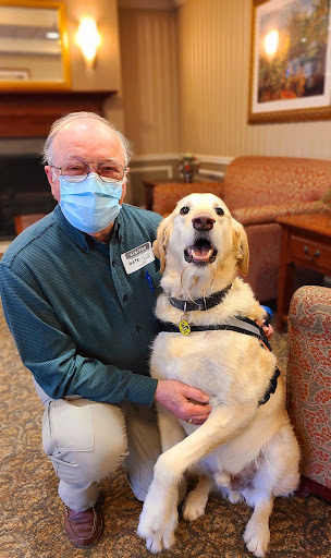 A smiling individual wearing a mask poses with a happy Golden Retriever in a cozy indoor setting.