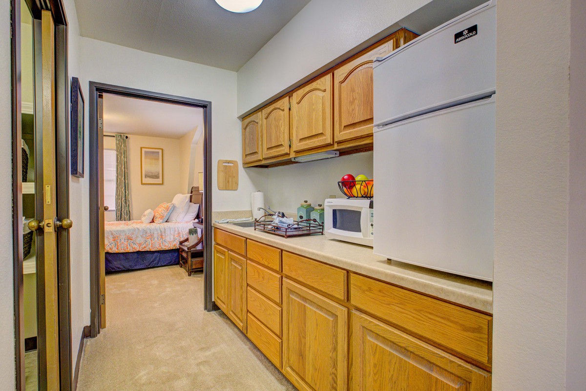 A compact kitchen area with wooden cabinets, a refrigerator, and a microwave, leading into a bedroom.