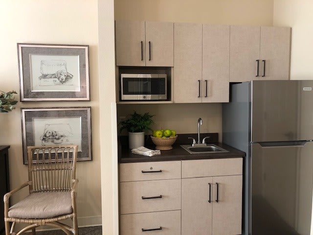 A compact modern kitchen with light cabinetry, featuring a microwave, refrigerator, and a small sink area.