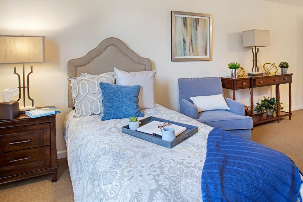 A cozy and well-decorated bedroom featuring a bed with patterned bedding, a blue throw blanket, and cushions. A tray is placed on the bed, and there is a comfortable chair along with a side table topped with a lamp.