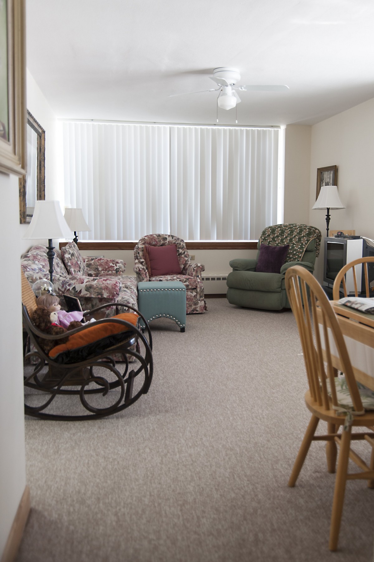 A cozy living room with various seating options, including a rocking chair and armchairs, complemented by a dining table and window blinds.