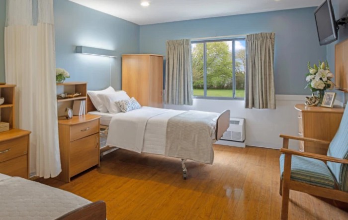 A clean and minimalist hospital room with a single bed, wooden floor, and large window providing natural light.