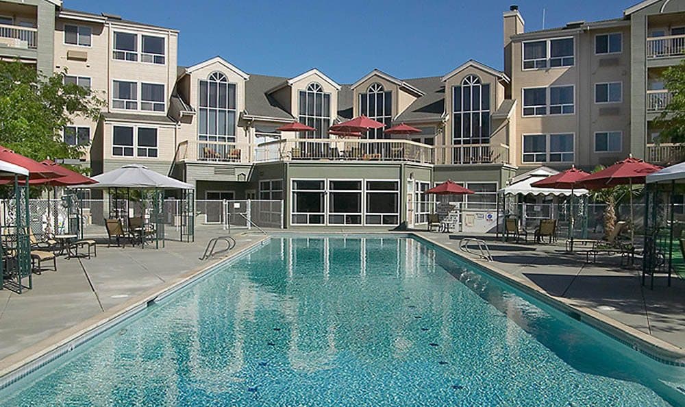 A spacious outdoor swimming pool in front of a modern apartment complex under a clear blue sky.