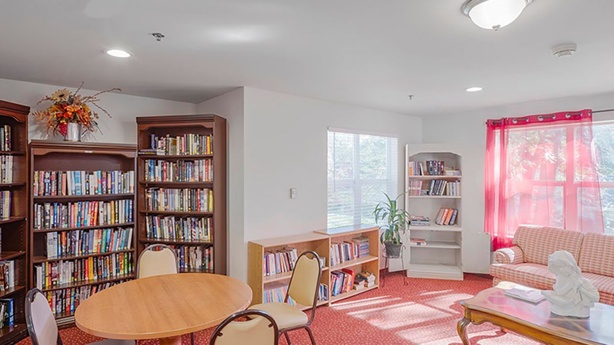 A cozy library room with bookshelves, tables, and chairs next to a window.