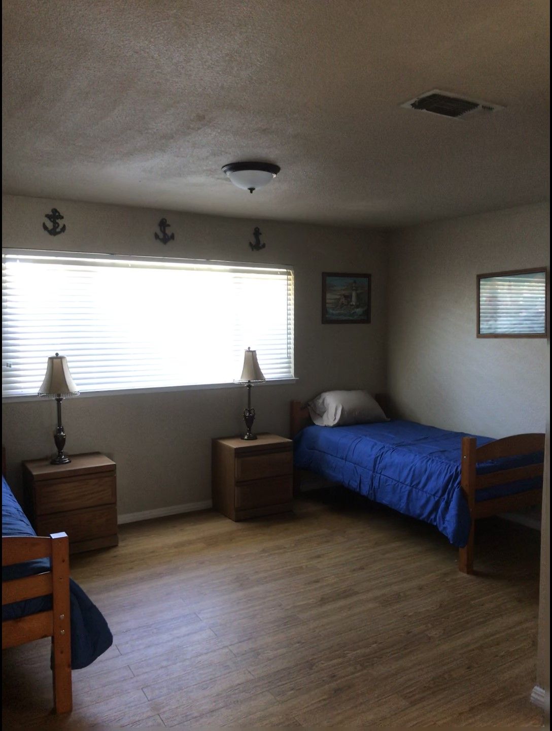 A simple bedroom with two single beds, each with blue bedding. The room features wooden floors, a large window, nightstands, and lamps.