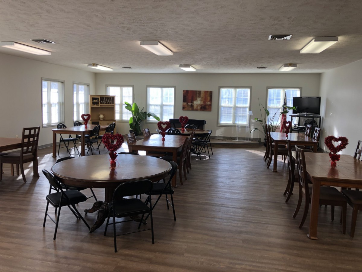 A spacious dining area with round tables, each adorned with red vases and surrounded by chairs, illuminated by natural light from large windows.