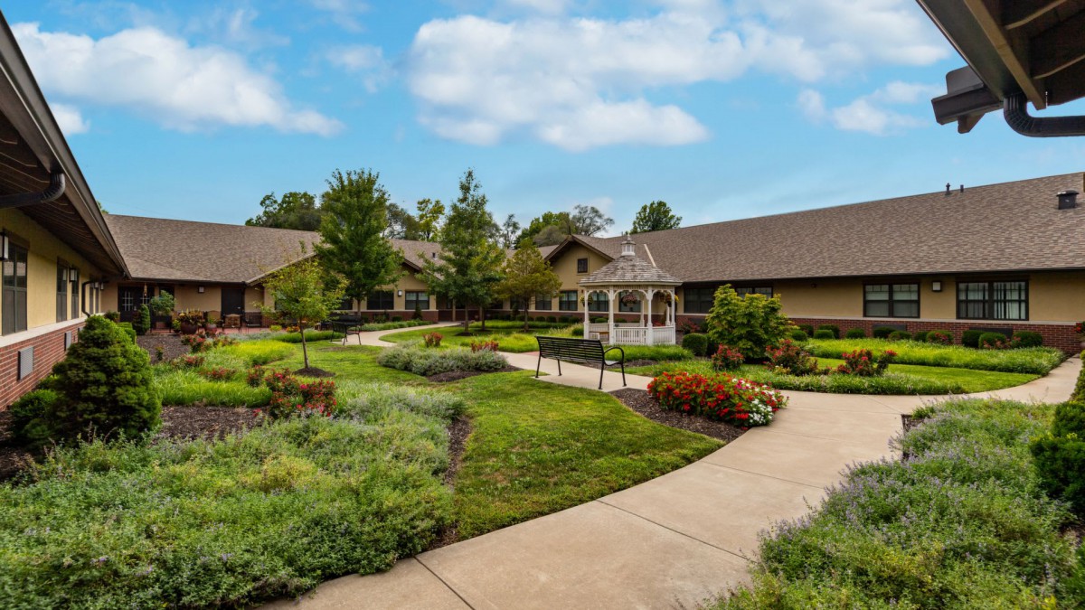 A serene garden with a pathway winding through lush greenery and leading to a gazebo, bordered by buildings under a blue sky.