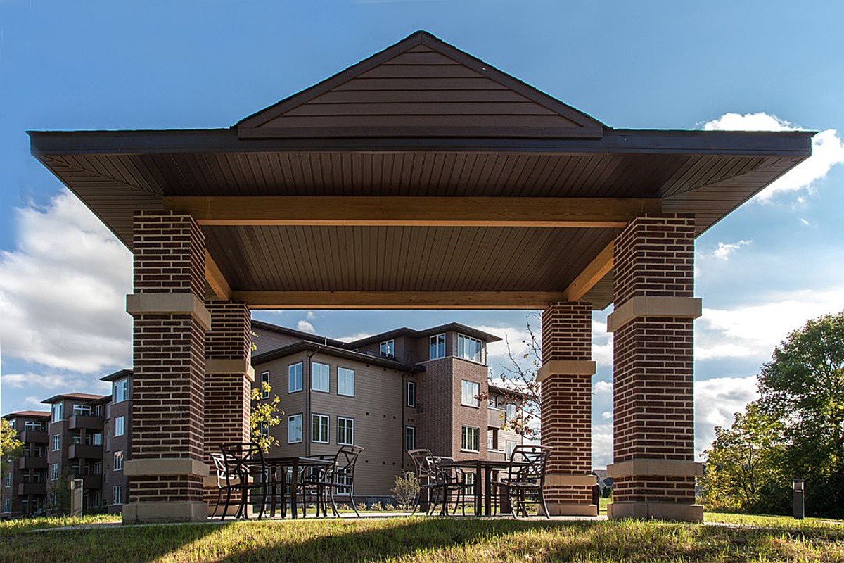 A brick gazebo frames a view of a residential building with outdoor tables and chairs on a sunny day.