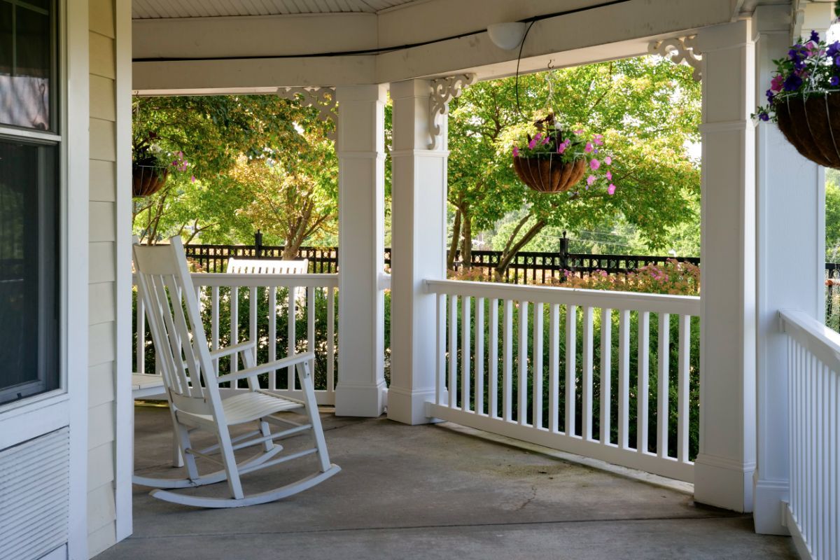 A serene porch with a white rocking chair and hanging plants overlooks a sunny garden.