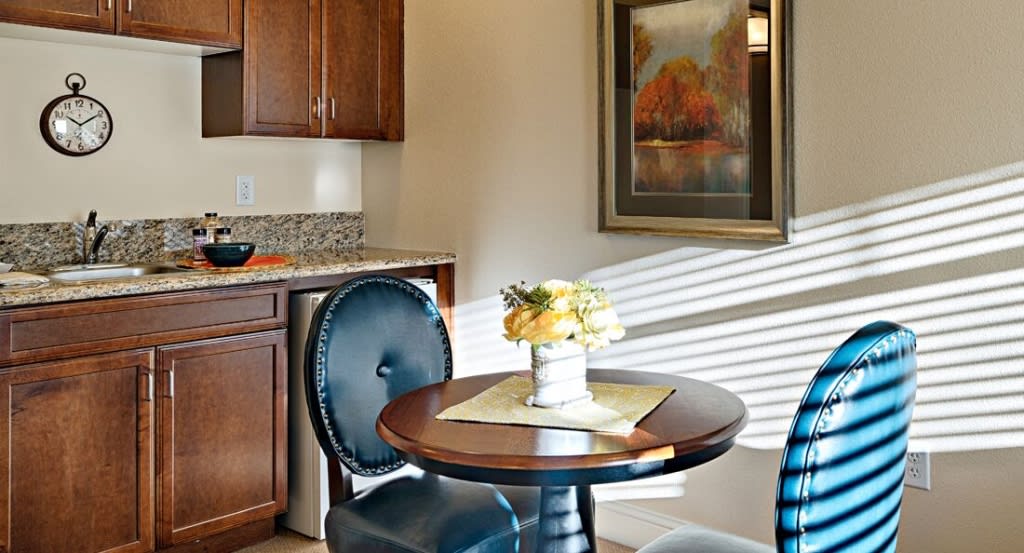 A cozy dining area with a round table, two chairs, and a flower arrangement, next to a kitchen.