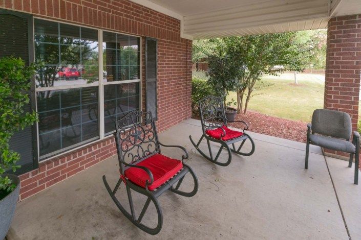 A cozy porch with two rocking chairs featuring red cushions, set against a brick wall and overlooking a garden.