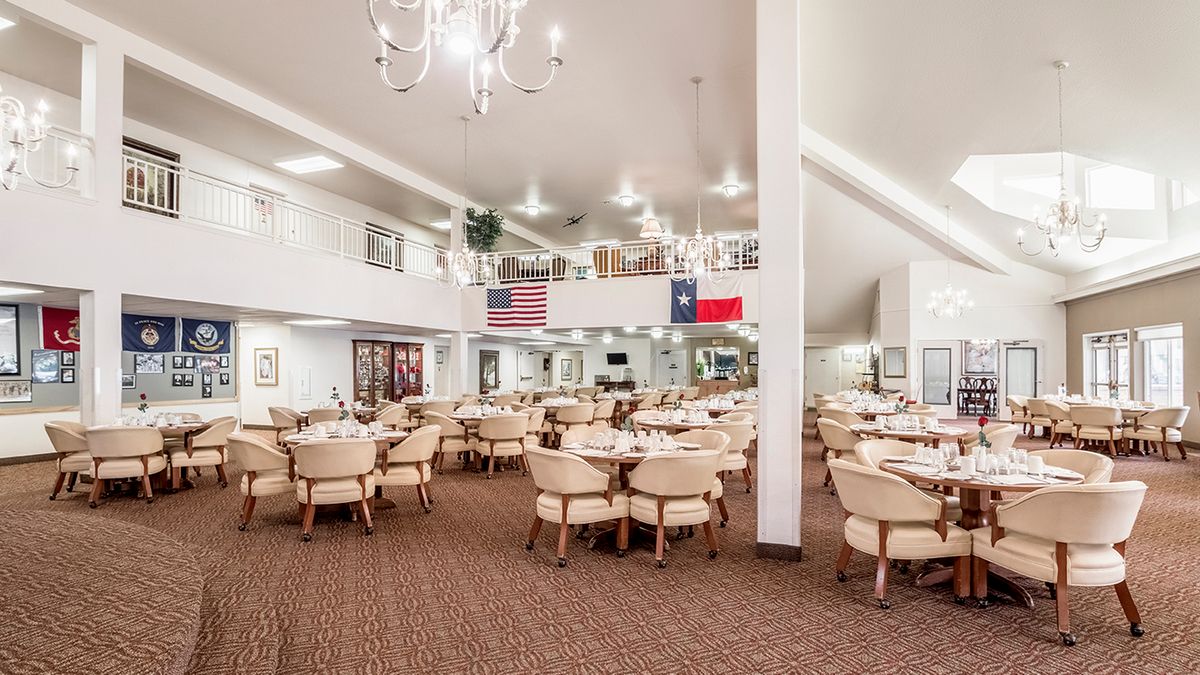 A spacious dining room with multiple tables and chairs, decorated with chandeliers and flags.