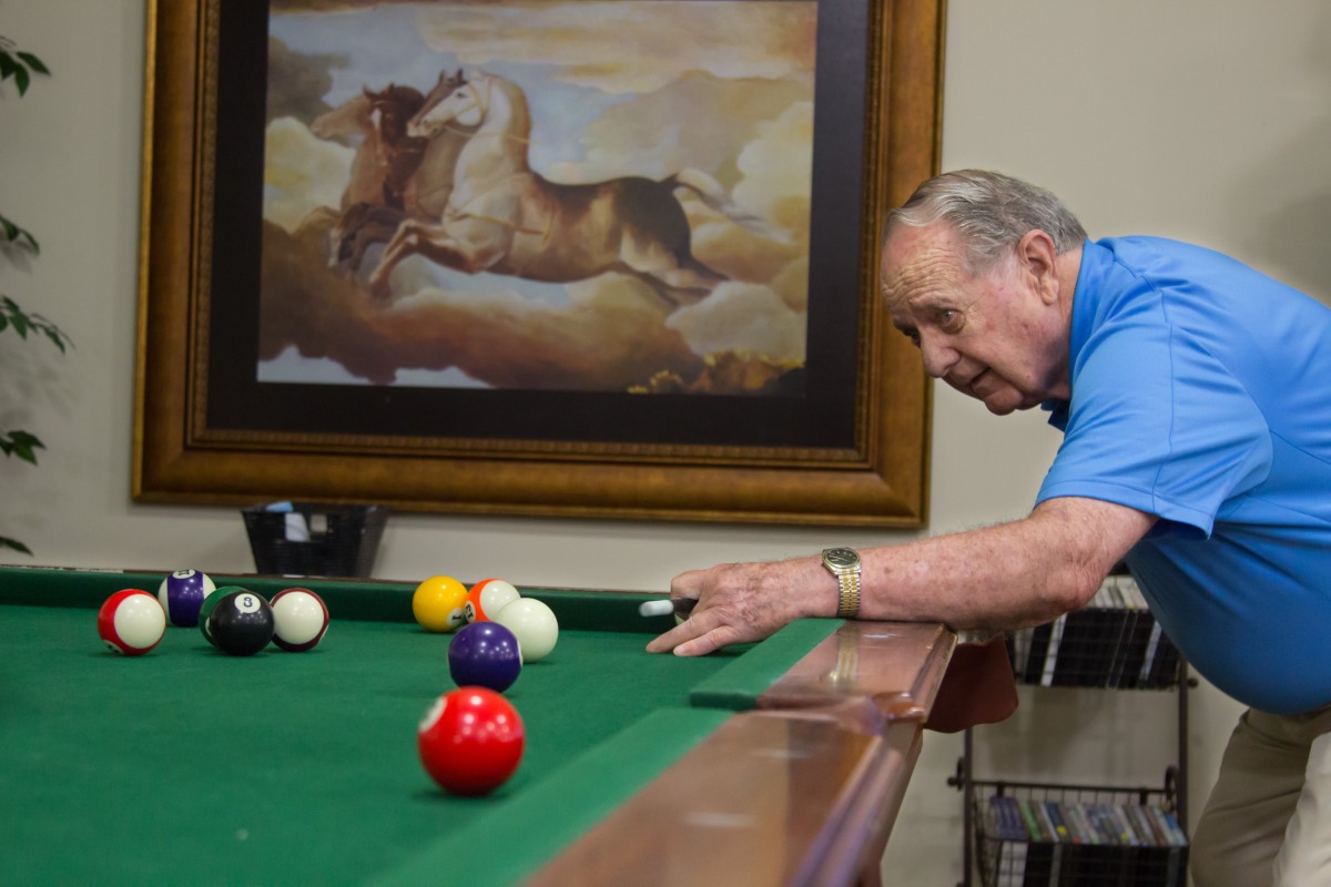 An elderly man playing billiards on a pool table with a painting in the background.