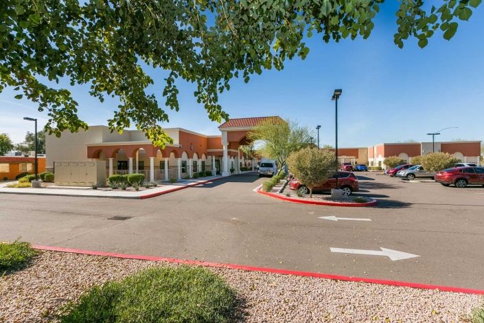 A view of a building entrance with a parking lot and landscaped surroundings on a clear day.