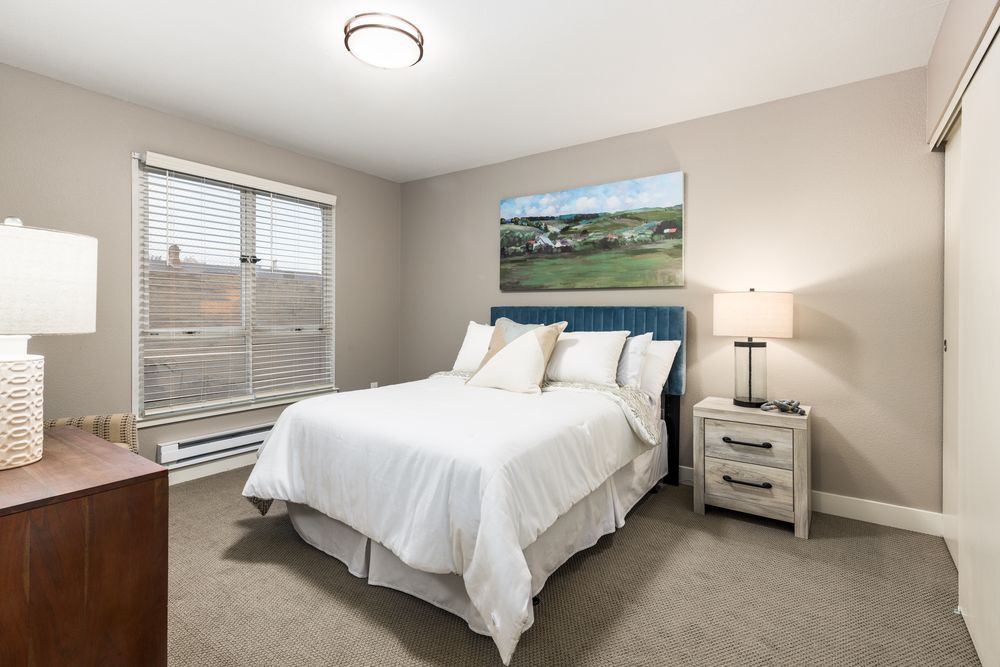 A cozy bedroom with a neatly made bed, a nightstand with a lamp, and a large window providing natural light.