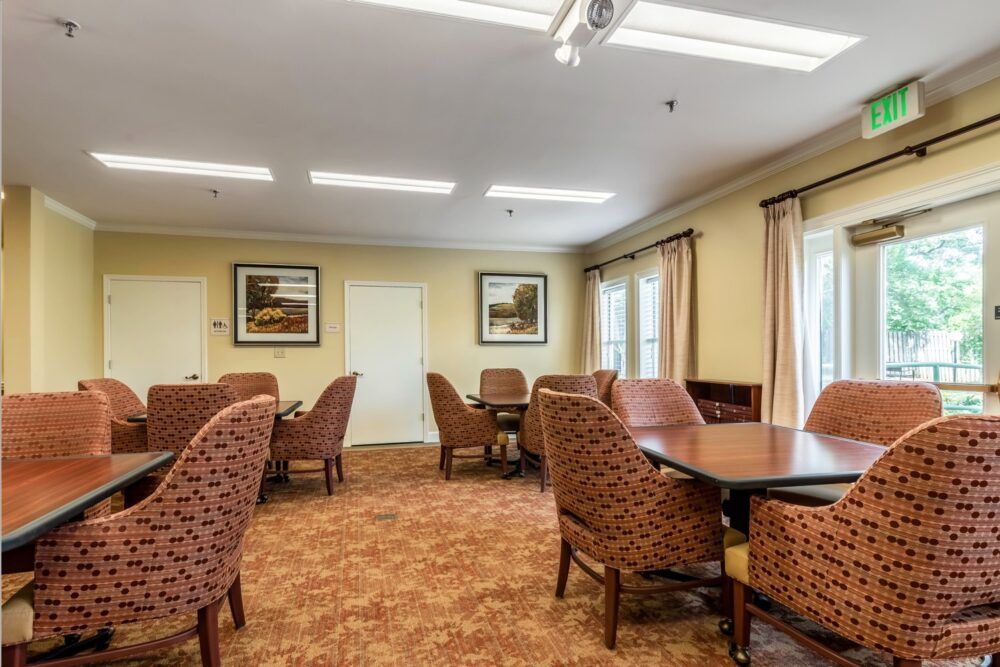 A cozy dining area with multiple tables and upholstered chairs, decorated with paintings and curtains.