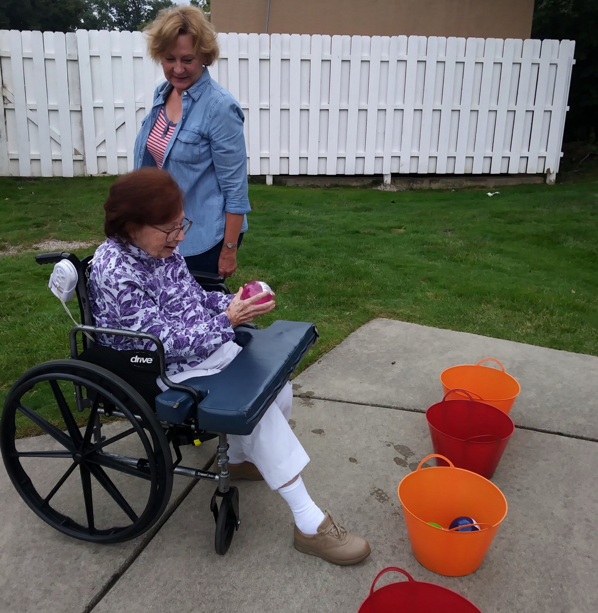 An elderly person in a wheelchair playing a game with buckets outdoors, accompanied by another person.