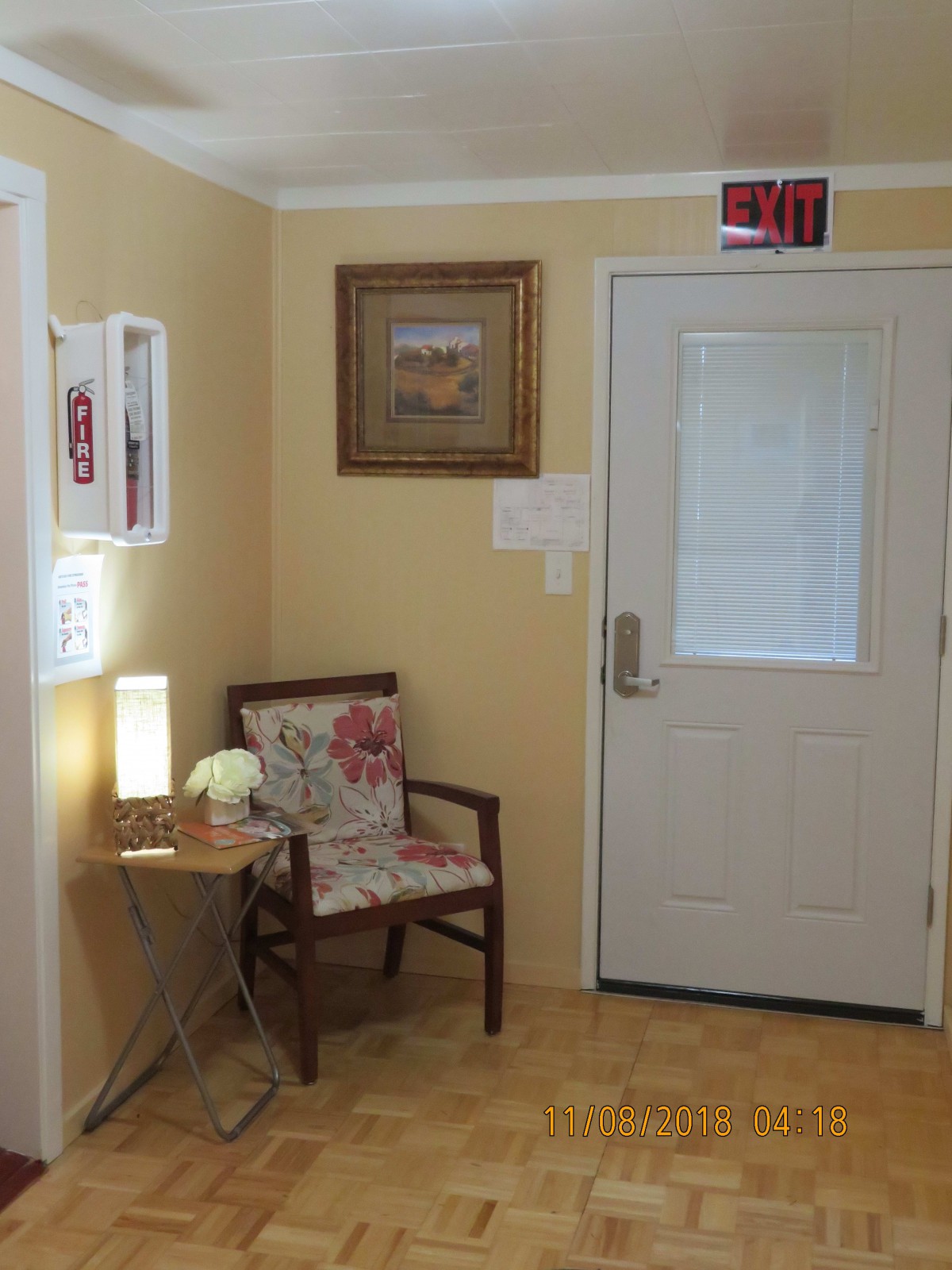 A cozy waiting area with a chair, side table, lamp, and art on the wall near an exit door.