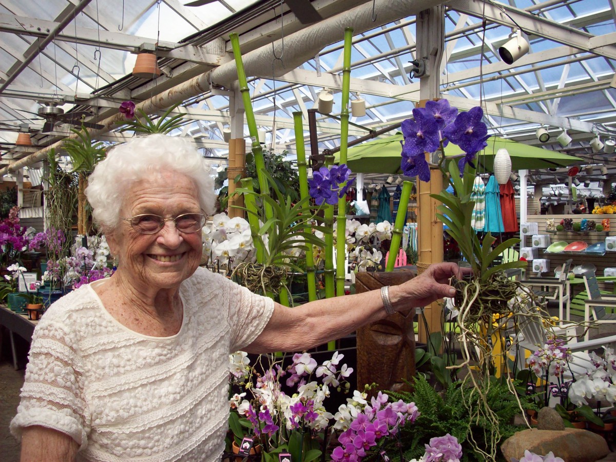 An elderly woman happily stands in a greenhouse surrounded by colorful flowers and plants.